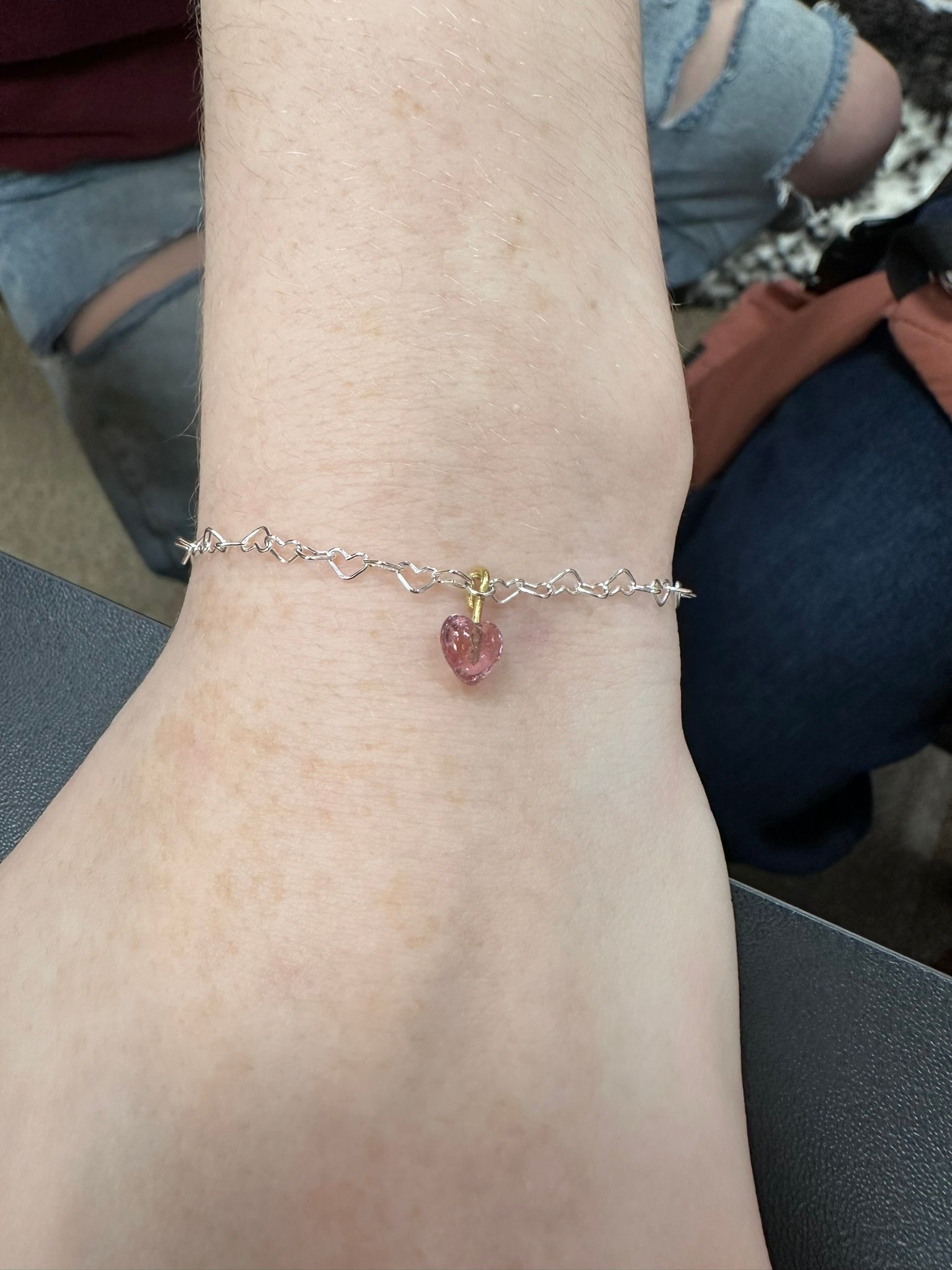 Wrist with a delicate silver bracelet featuring a small, heart-shaped pink gem.