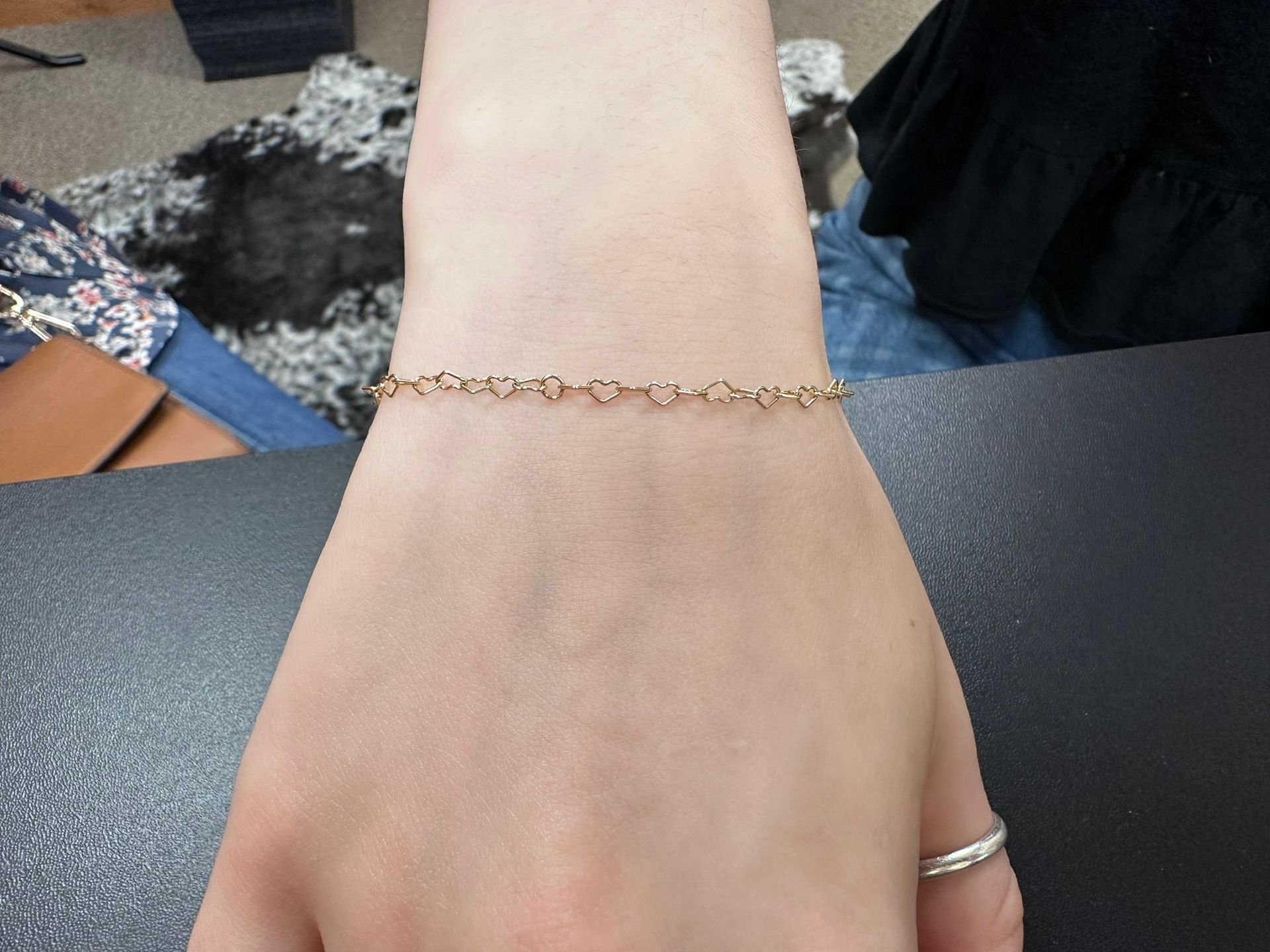 Person's arm with a gold bracelet featuring small heart-shaped links. A silver ring is on the finger.