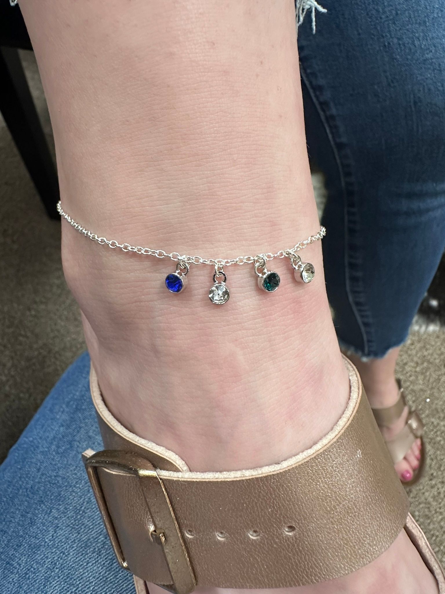 Anklet with colorful charms on a person's ankle, wearing gold sandals and jeans.