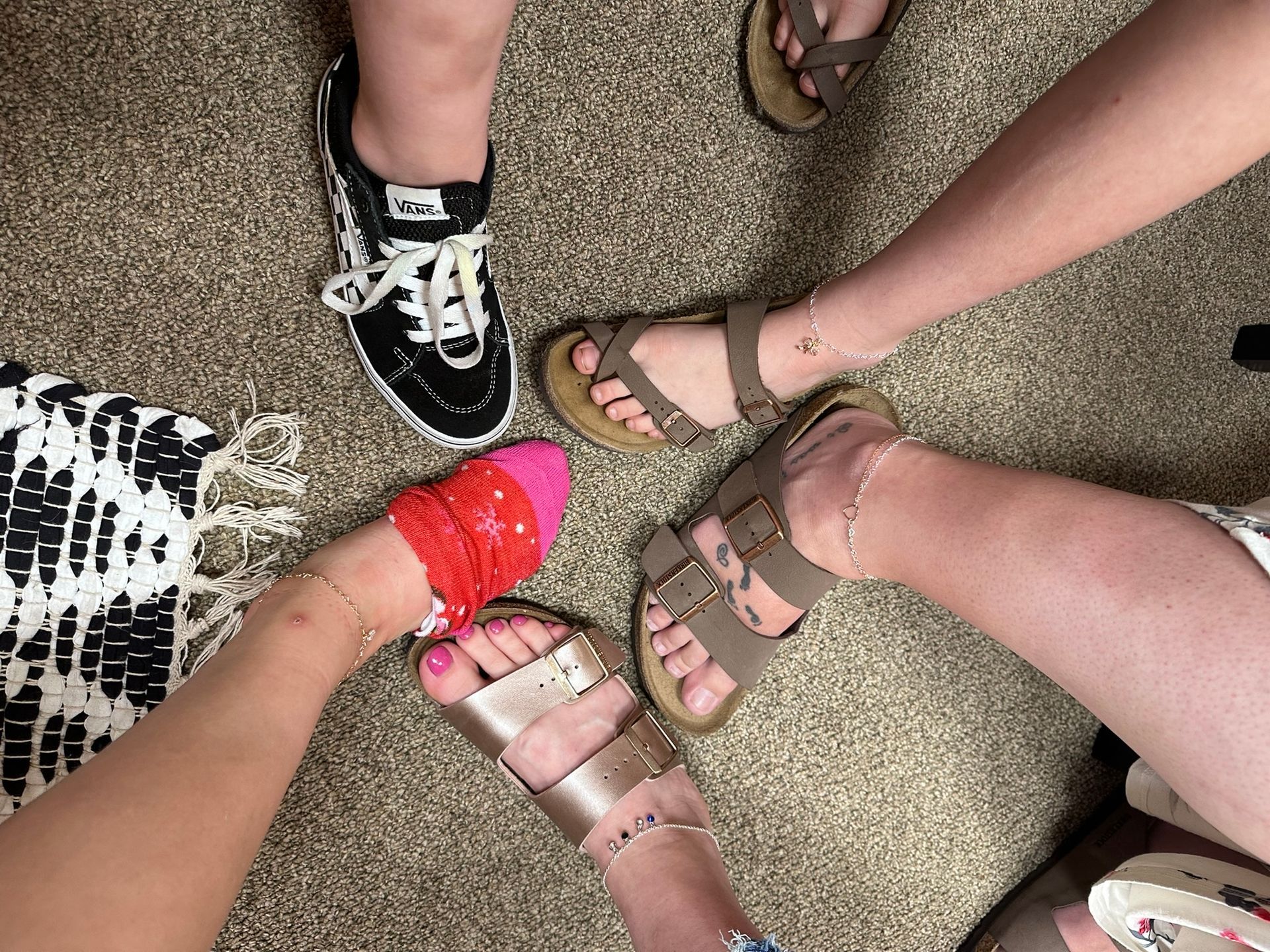 Five people's feet in a circle, wearing sneakers, sandals, and a sock. The feet have anklets.