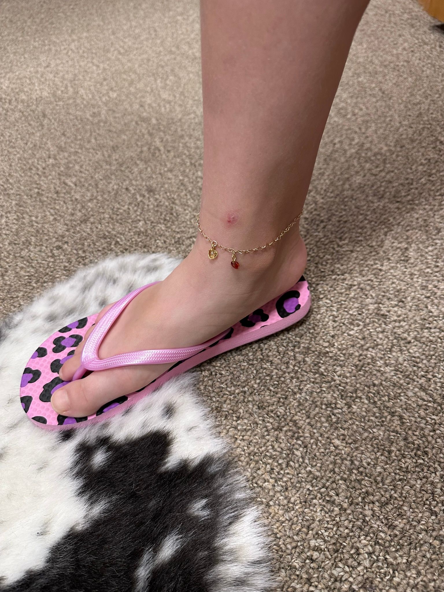 A person's foot in a pink leopard print flip-flop with a gold anklet on a rug.