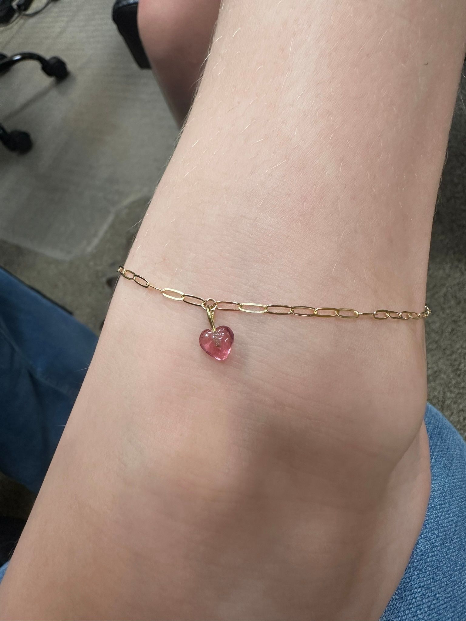 Anklet with heart pendant on ankle. Gold chain, pink charm, blue jeans visible.