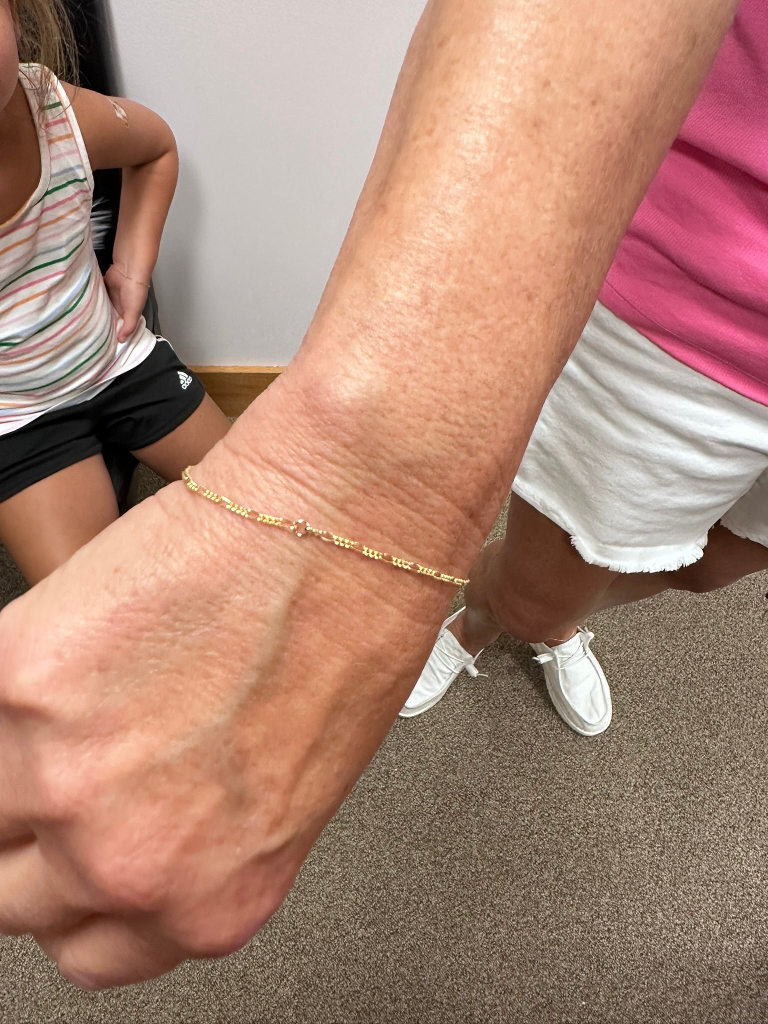 Person's wrist with gold chain bracelet. Another person stands nearby.