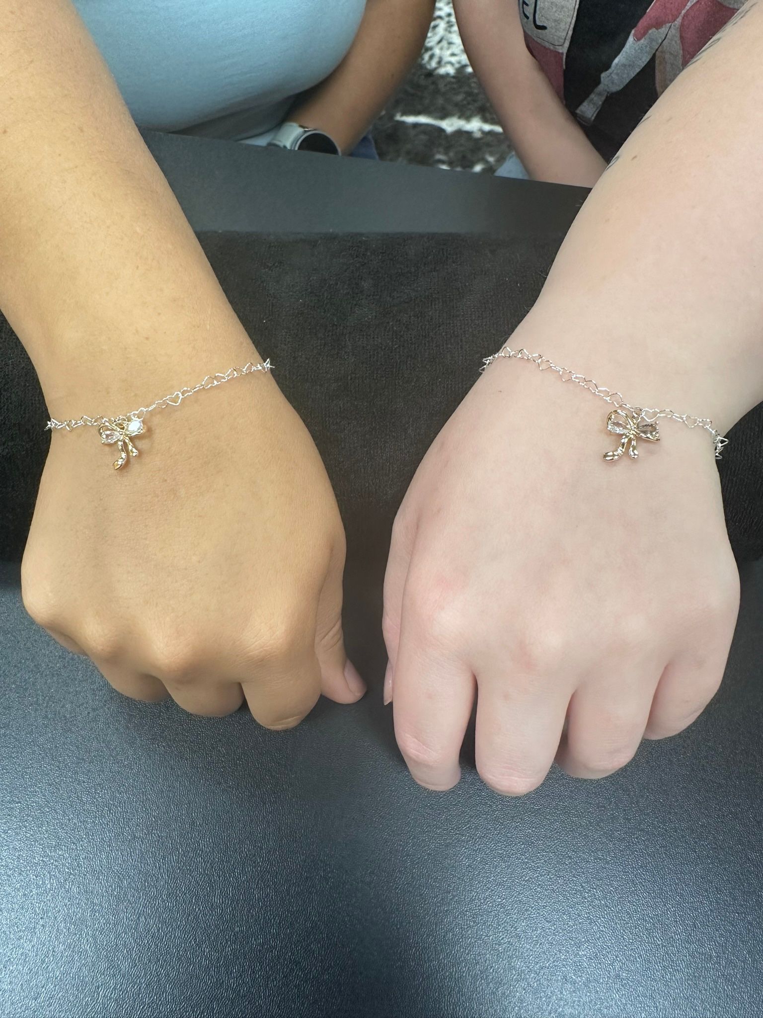 Two arms side-by-side on a dark surface, each with a silver bracelet and a charm. One arm is tan, the other is fair.