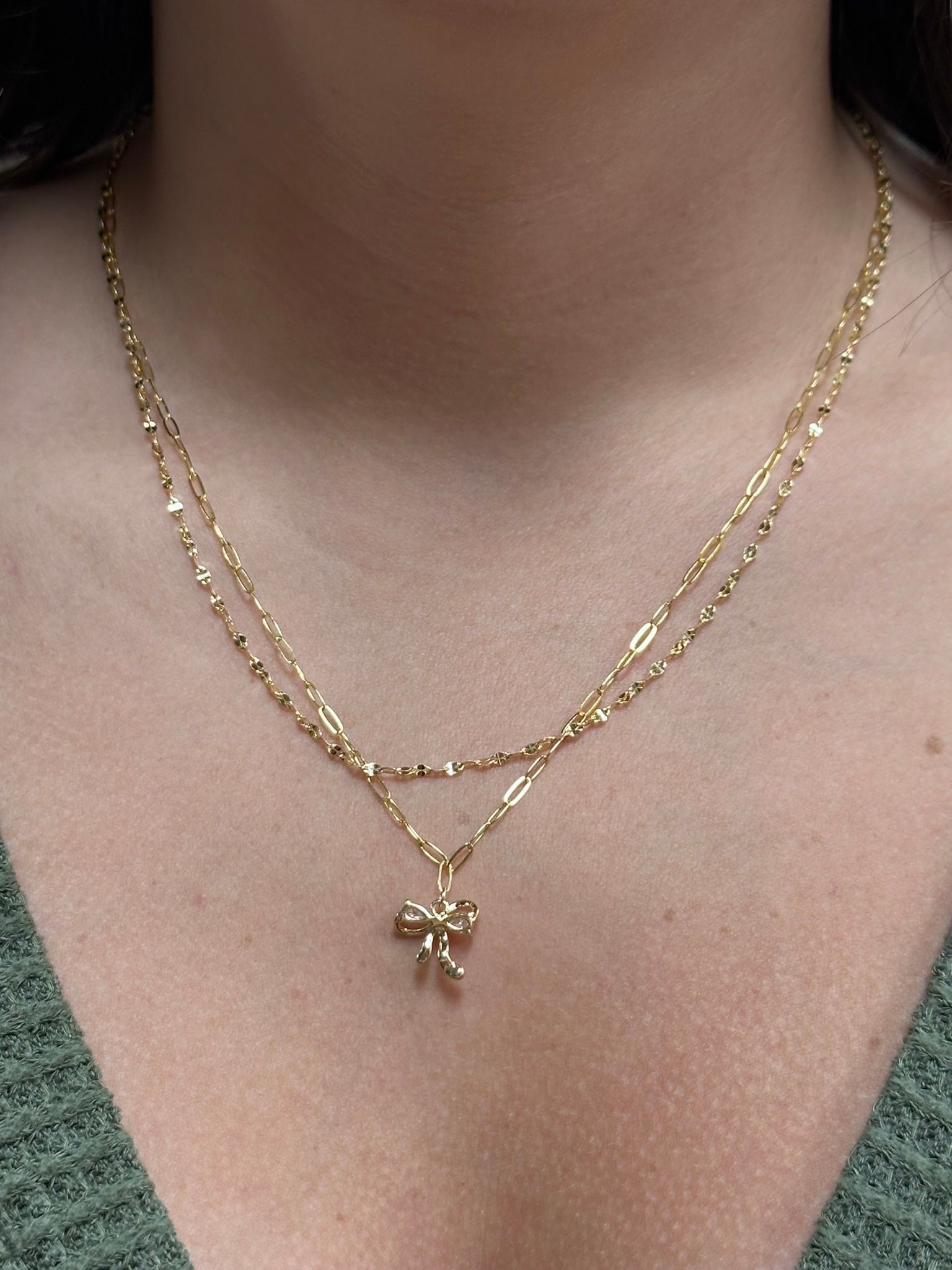 Gold layered necklace with small bow pendant on a person's neck.