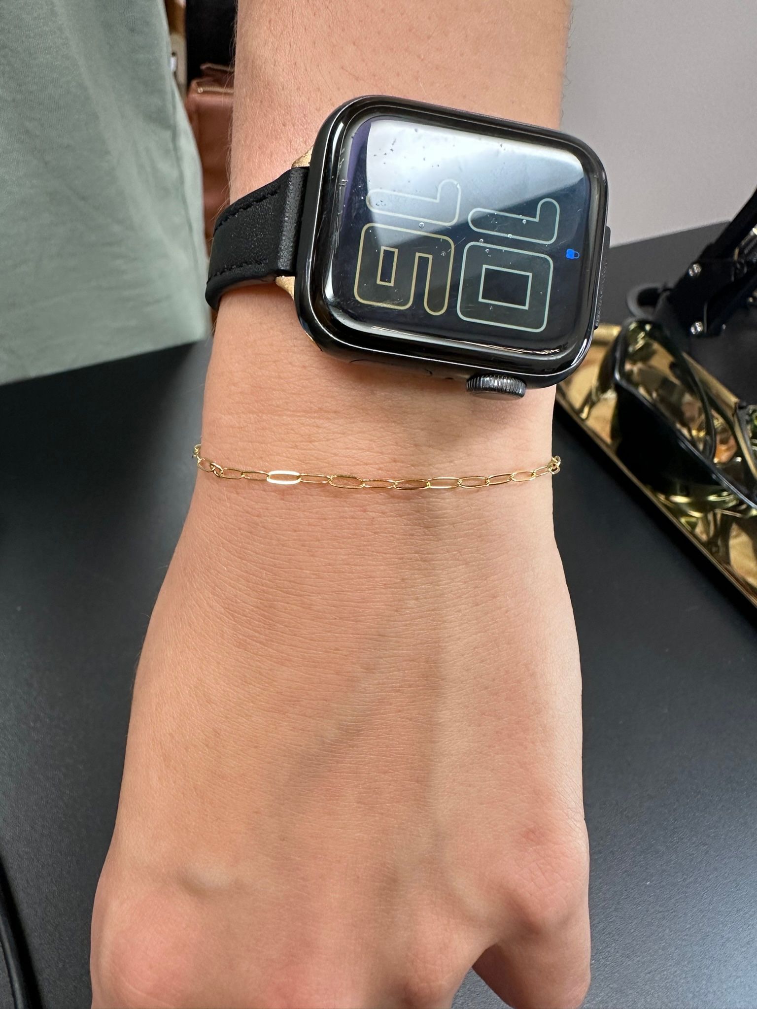 Wrist with a gold chain bracelet and a black smartwatch with a custom face and a leather strap.