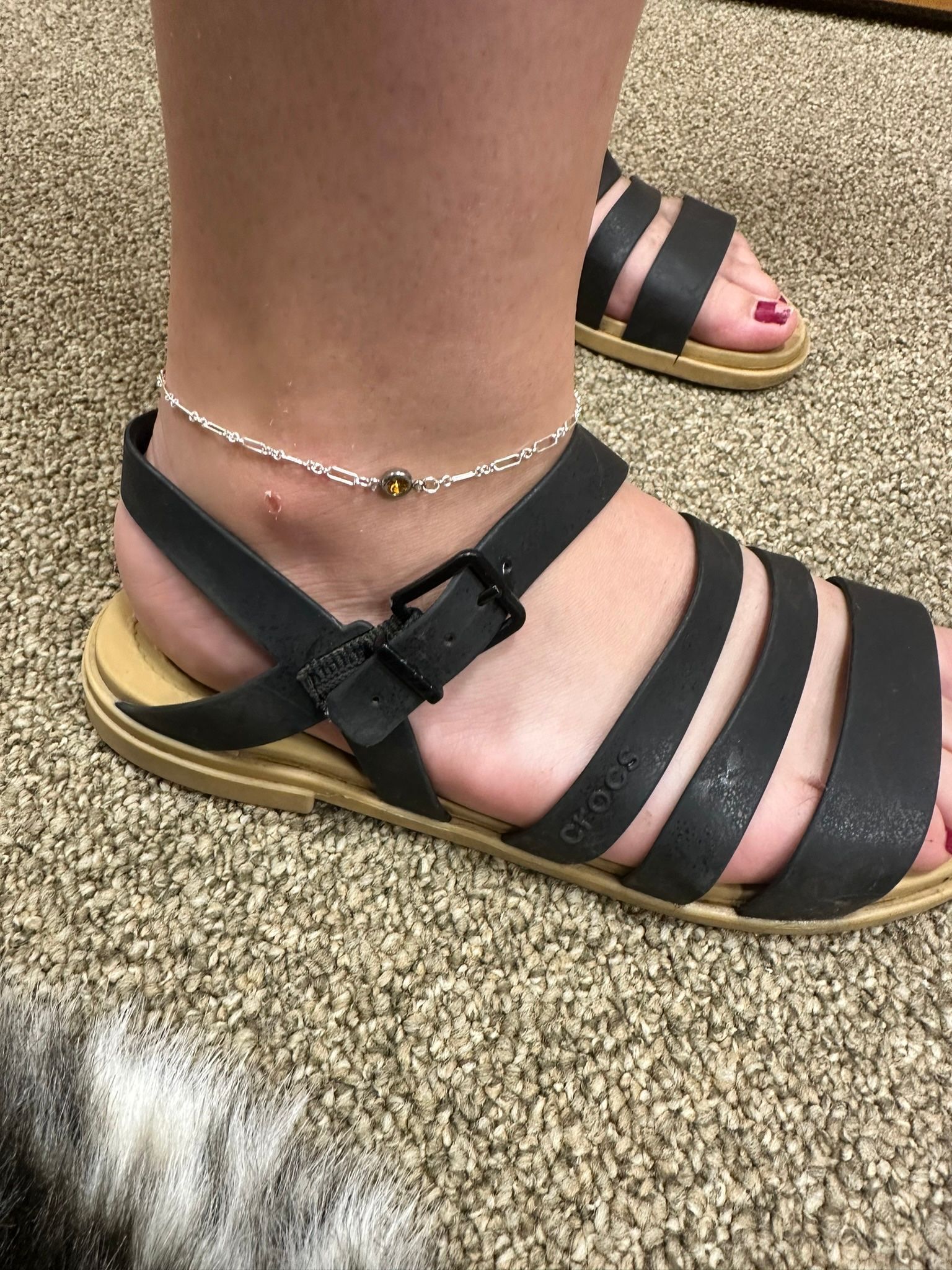 Person's foot in black sandals with a buckle and silver anklet, standing on beige carpet.