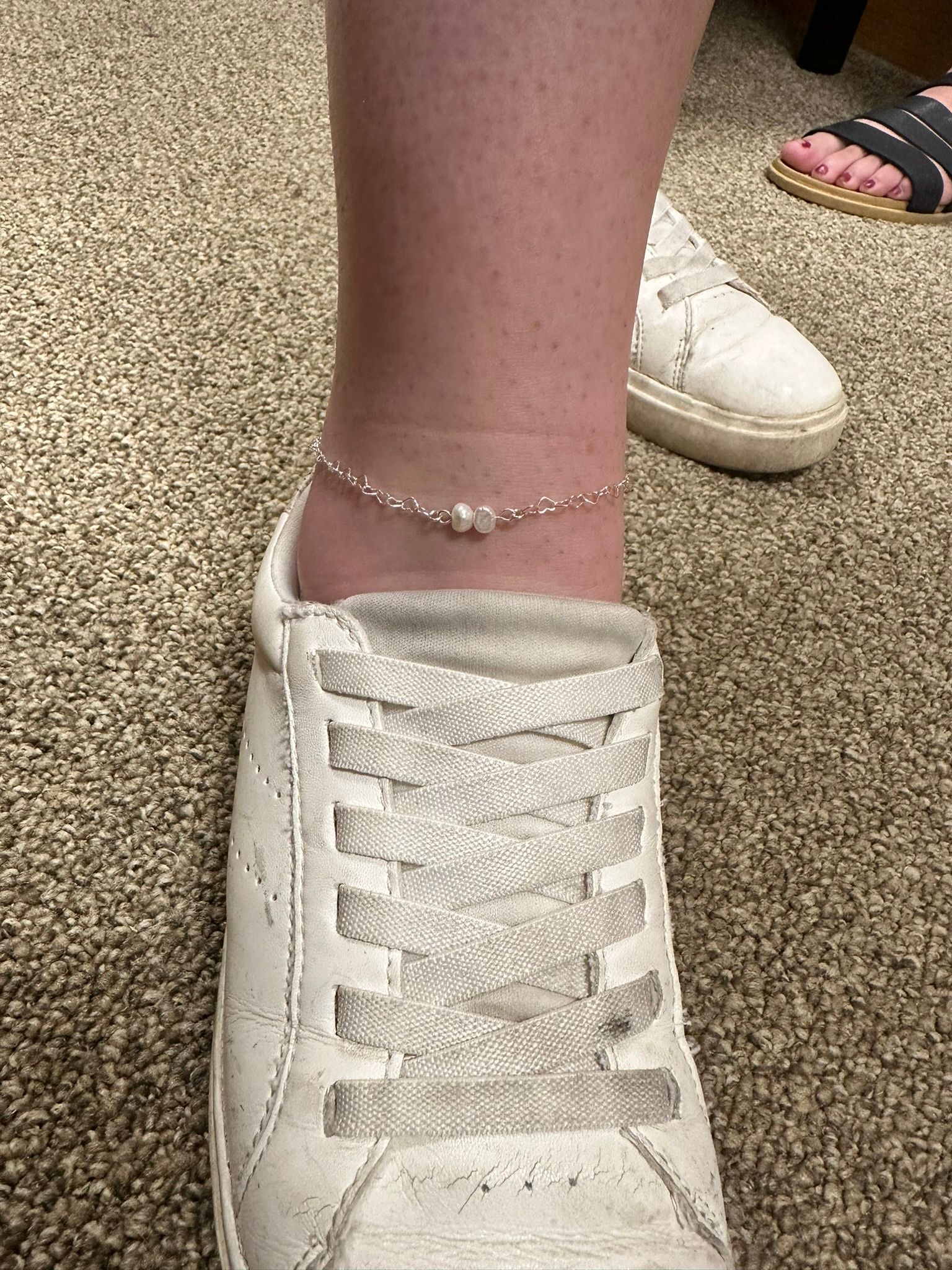 Ankle with silver anklet, white sneaker. Brown carpet in the background. Another foot in sandals visible.