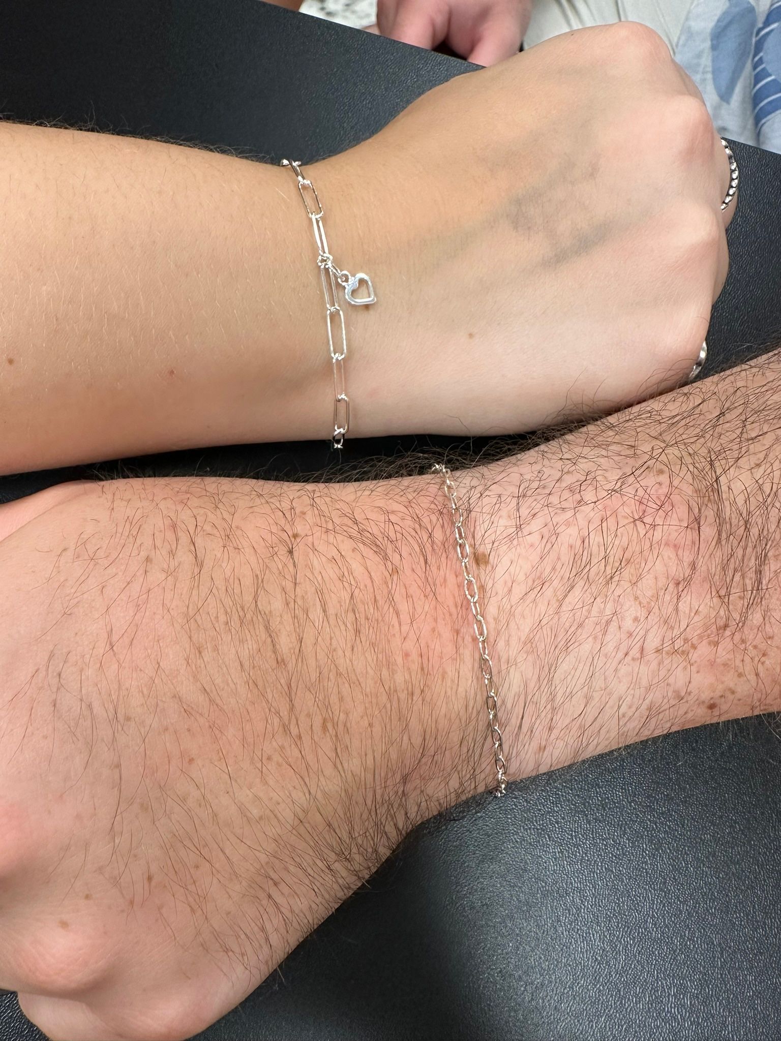 Two wrists with silver bracelets, one with a charm. One wrist is fair-skinned, the other has dark hair.