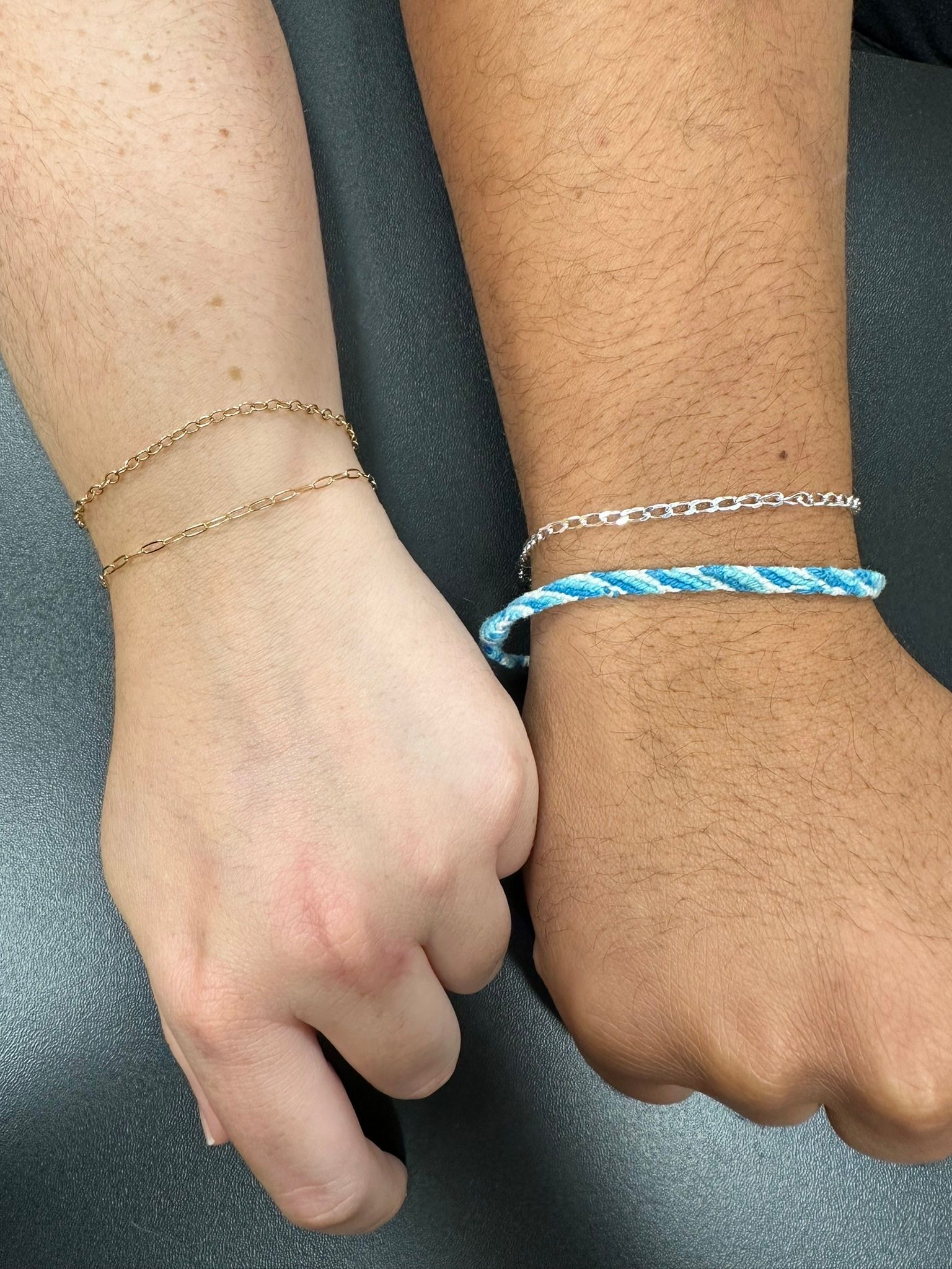 Two wrists with bracelets side by side; one with gold chains, the other with silver and blue woven bracelet.