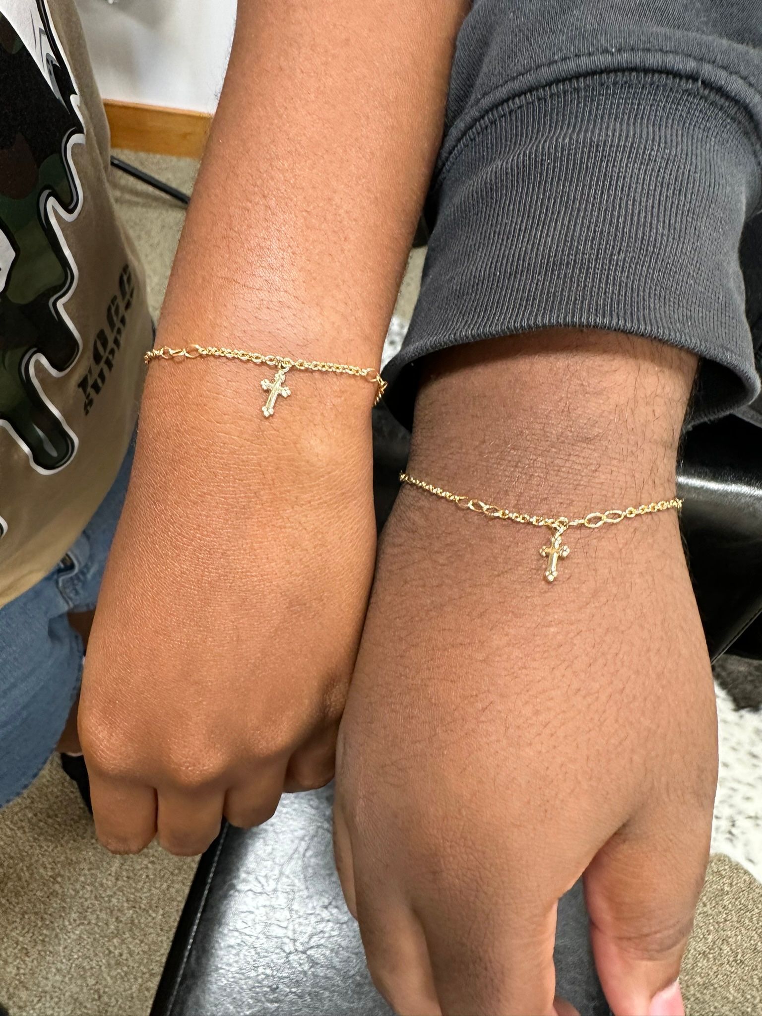 Two arms, each wearing a gold bracelet with a small charm, possibly crosses or letter “F”.