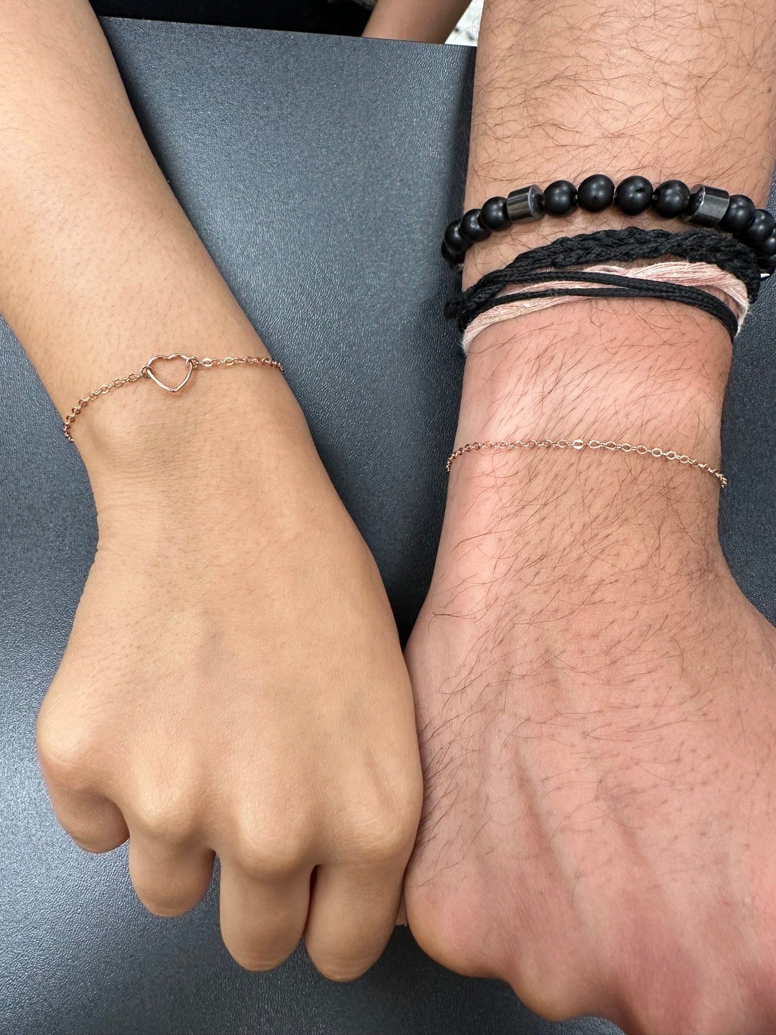 Two wrists, touching, with bracelets. One has a heart charm, the other has multiple beaded bracelets.