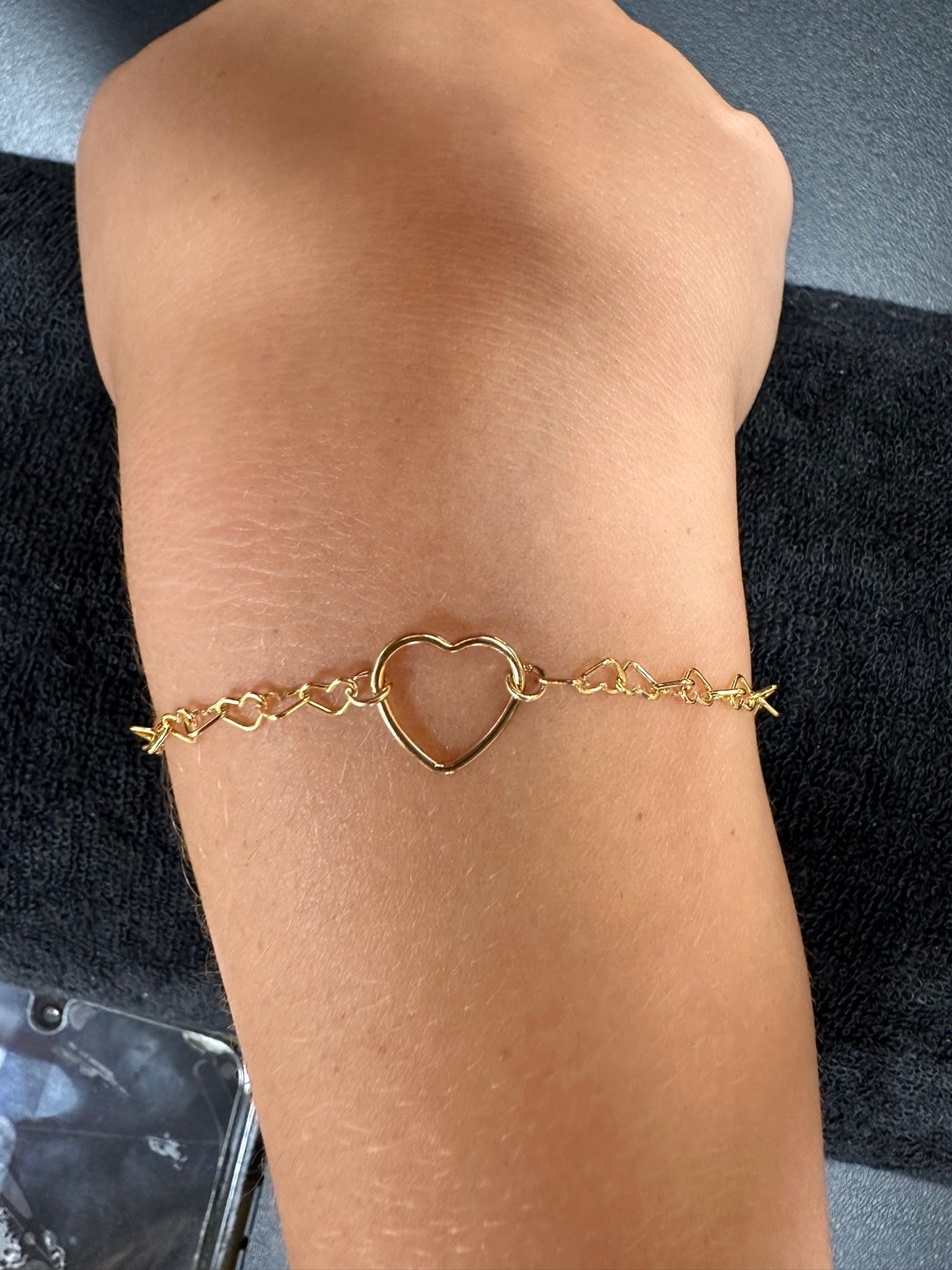 Gold heart-shaped bracelet on a wrist against a dark background.