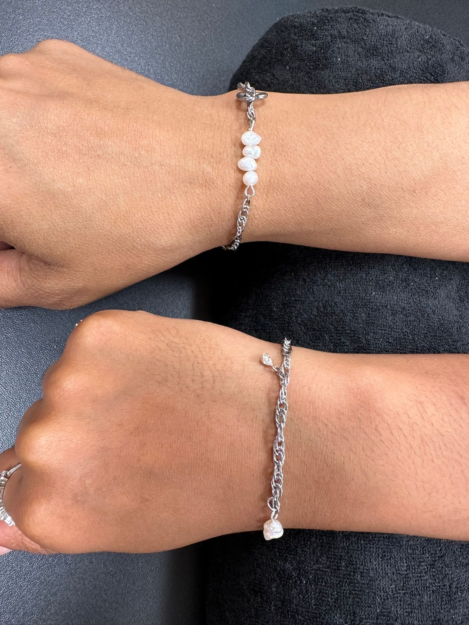 Two wrists with silver bracelets, one with white stones, resting on a gray surface.
