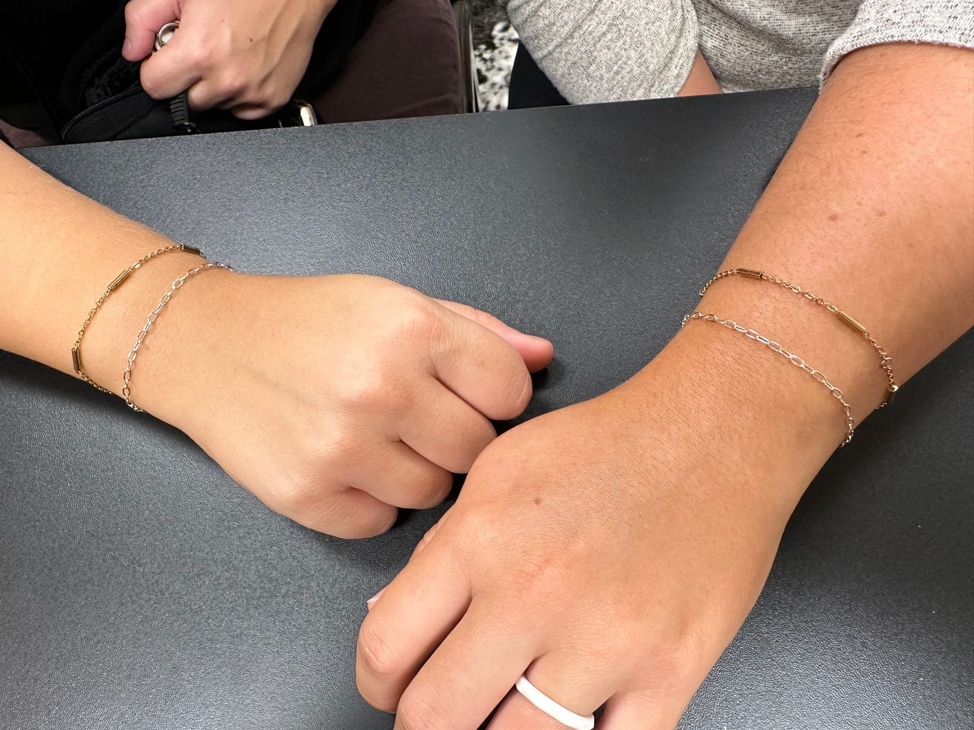 Two wrists, each wearing a gold bracelet, rest on a dark surface.