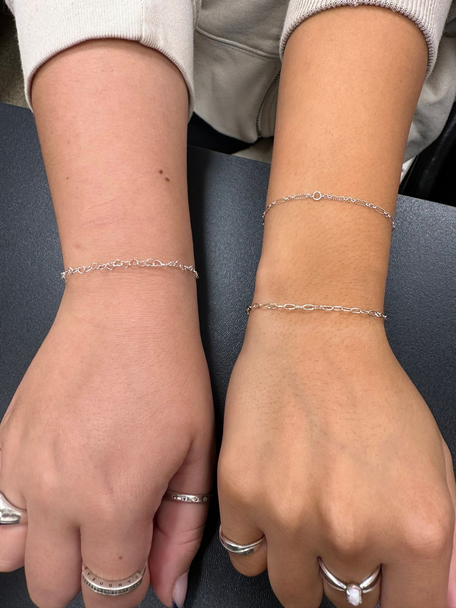 Two arms, one lighter, one tanned, each with silver bracelets and rings, resting on a surface.