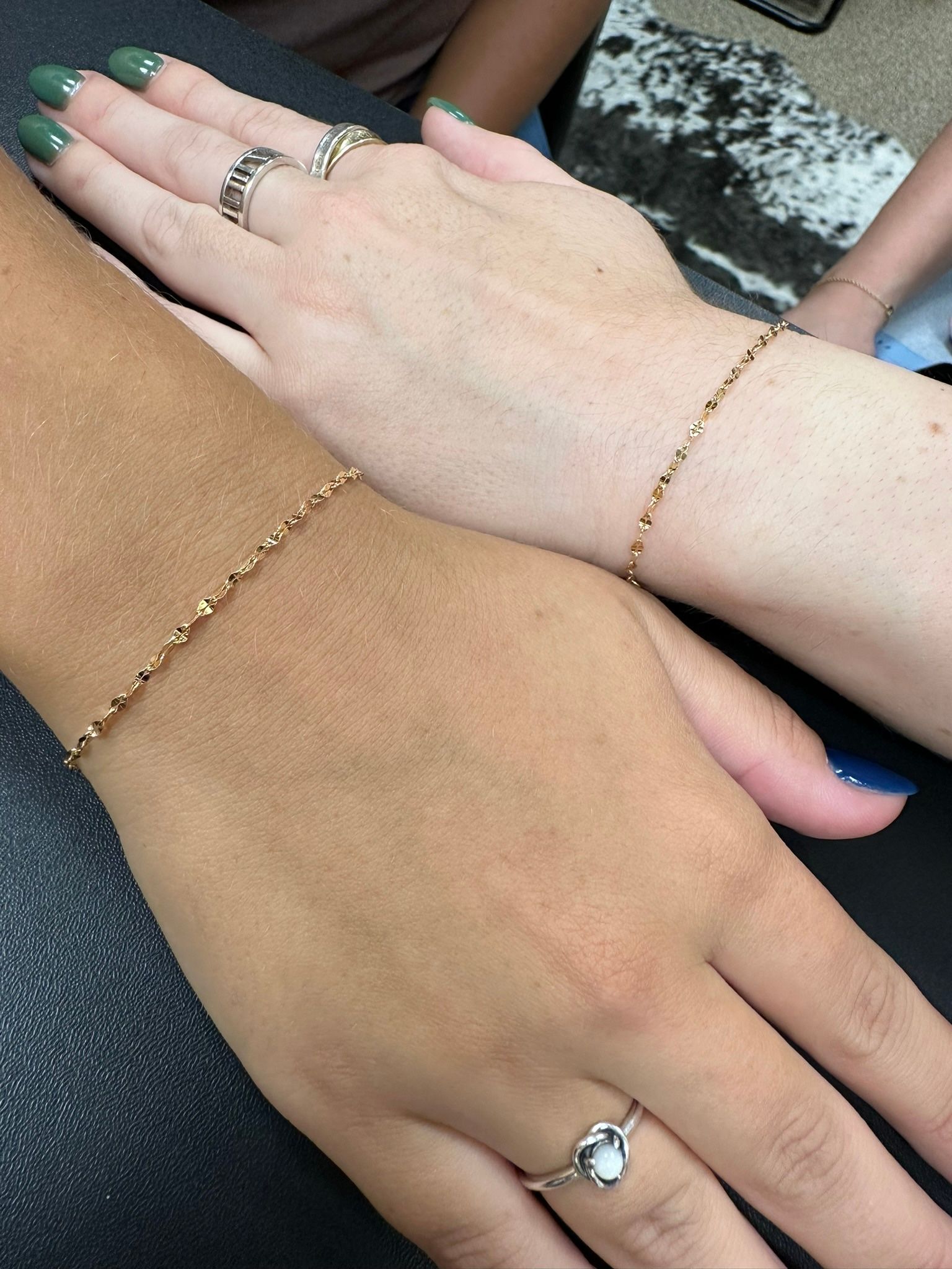 Two hands with matching gold bracelets, one darker and one lighter, side-by-side on a dark surface.