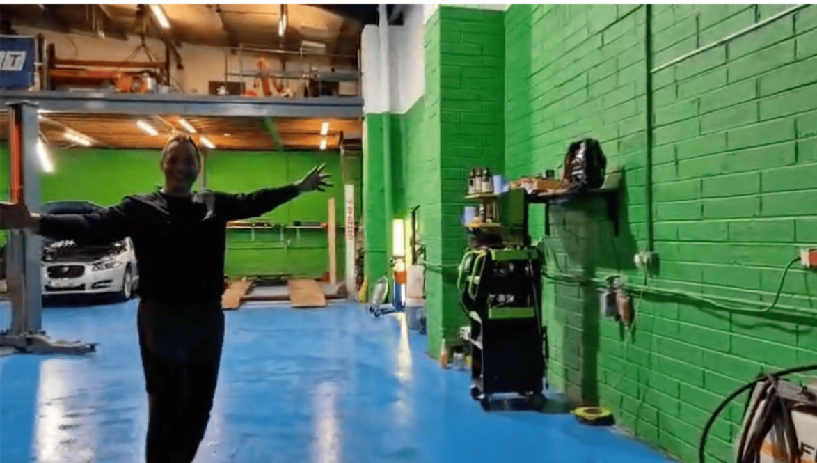 A person with arms outstretched stands in an auto repair shop with bright green walls and a blue floor.
