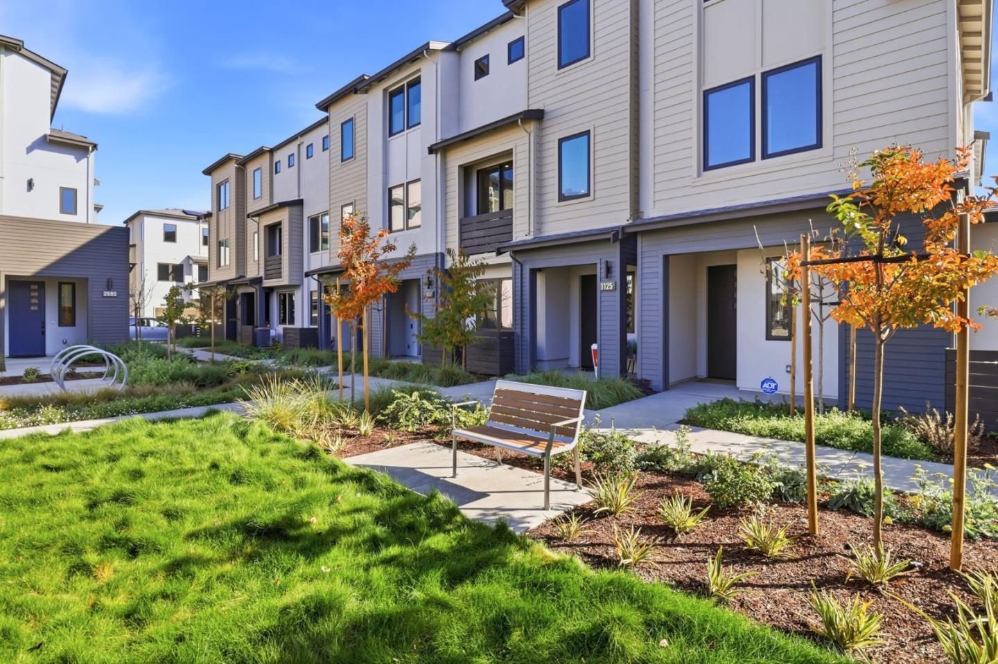 A modern apartment complex exterior with a grassy courtyard, a wooden bench, and small trees under a clear blue sky.