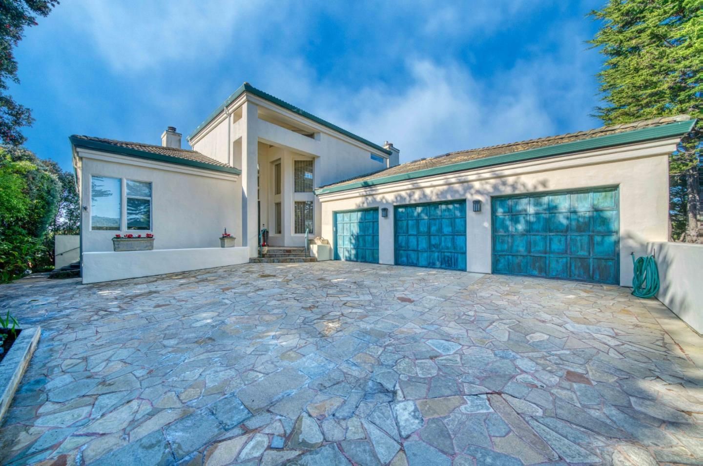 A two-story stucco house with a stone driveway, teal-colored garage doors, and a tiled roof under a blue, cloudy sky.