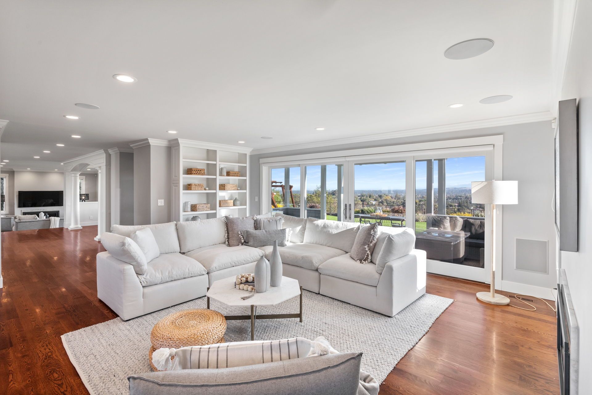 A bright, modern living room with a large white sectional sofa, wooden floors, and expansive glass doors overlooking a view.