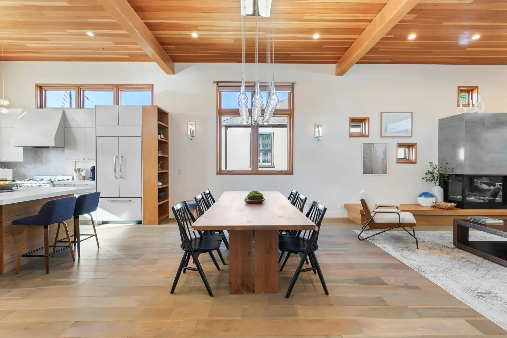 A bright, open-plan dining and kitchen area with light wood ceilings, hardwood floors, a long wooden table, and dark chairs.