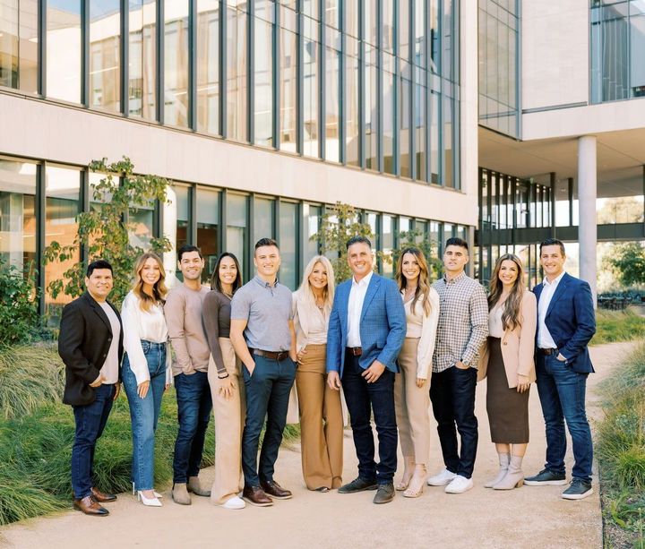 A group of eleven colleagues at Guide Real Estate stand together and smile in front of a modern glass office building.