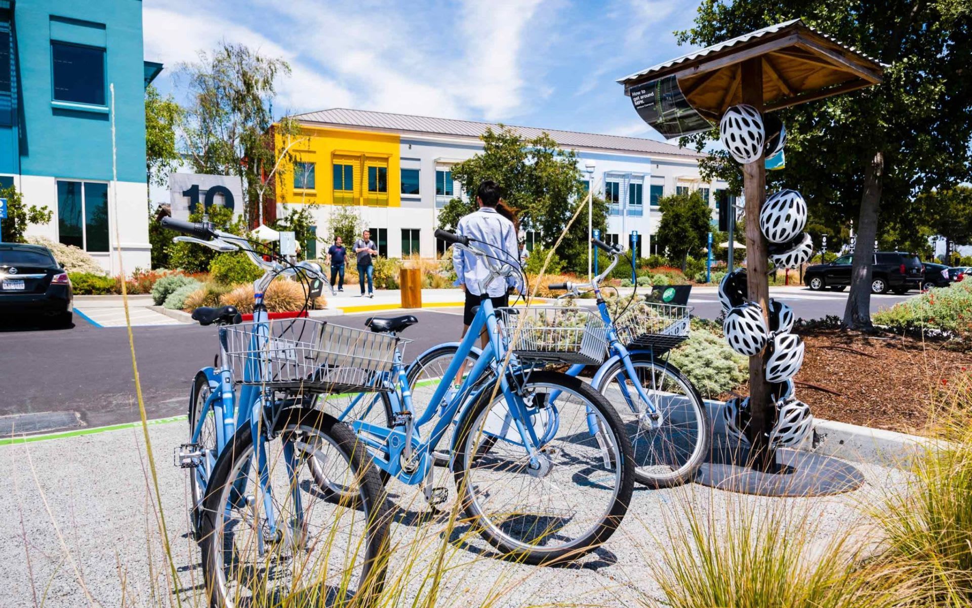 Two light blue bicycles are parked in front of a bike rack holding multiple white helmets on a sunny day near office buildings.