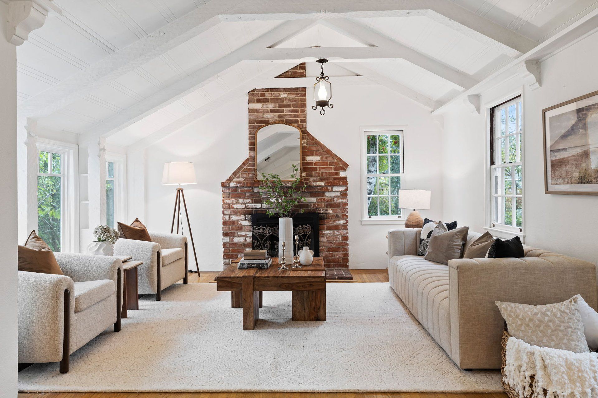 A bright, airy living room with a vaulted ceiling, exposed brick fireplace, neutral sofa, and two armchairs on a rug.