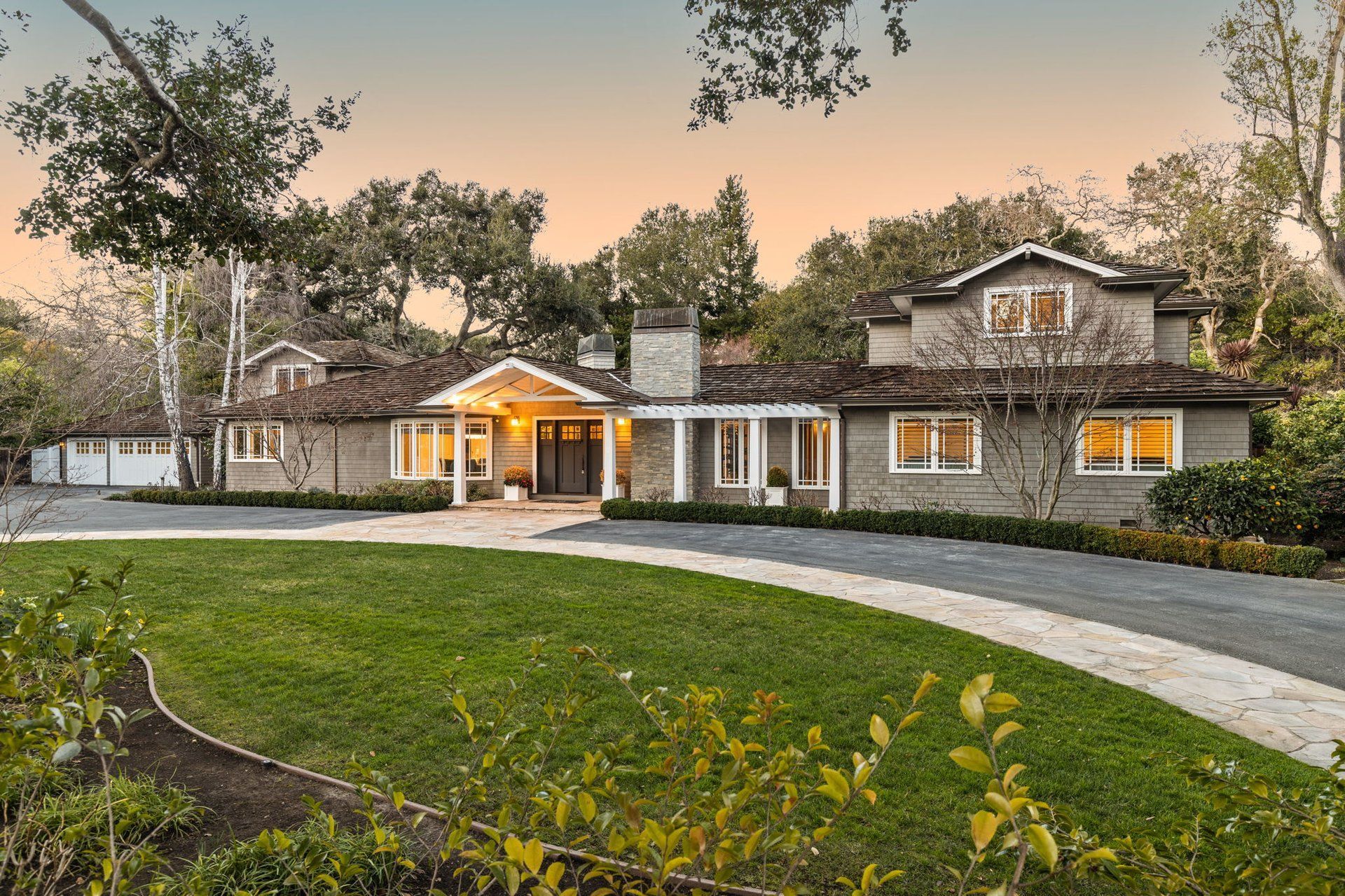 A sprawling, single-story light gray home with a stone chimney and wood-shingled roof, set behind a curved stone driveway.