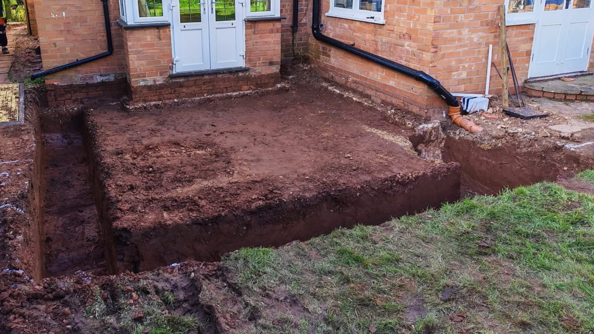 A backyard construction site shows a rectangular patch of excavated soil prepared for a foundation next to a brick house.