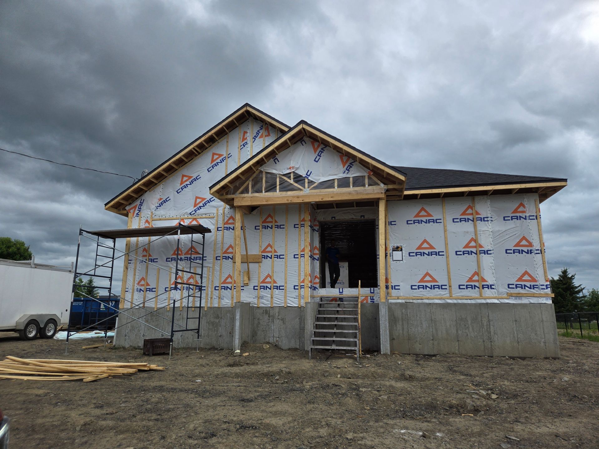 Maison en construction avec fondations en béton, recouverte d'une bâche résistante aux intempéries. Ciel sombre.