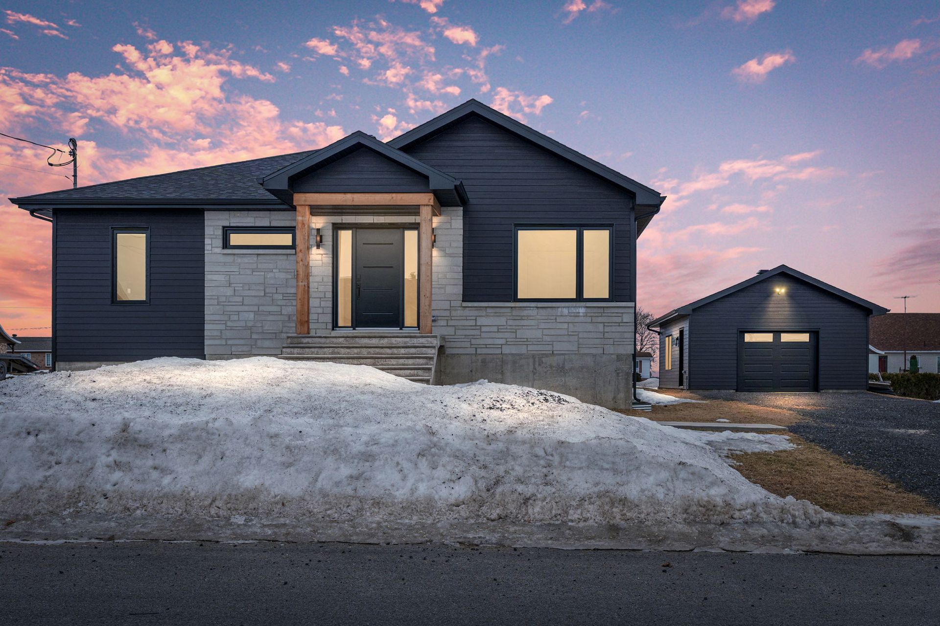 Maison moderne avec bardage sombre et façade en pierre, cour enneigée, garage, ciel crépusculaire.