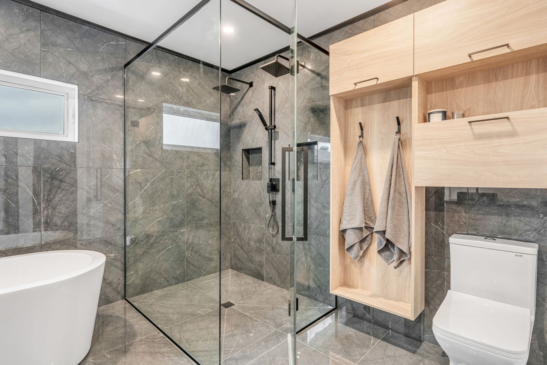 Salle de bains moderne avec carrelage gris, douche en verre, rangements en bois et toilettes blanches.