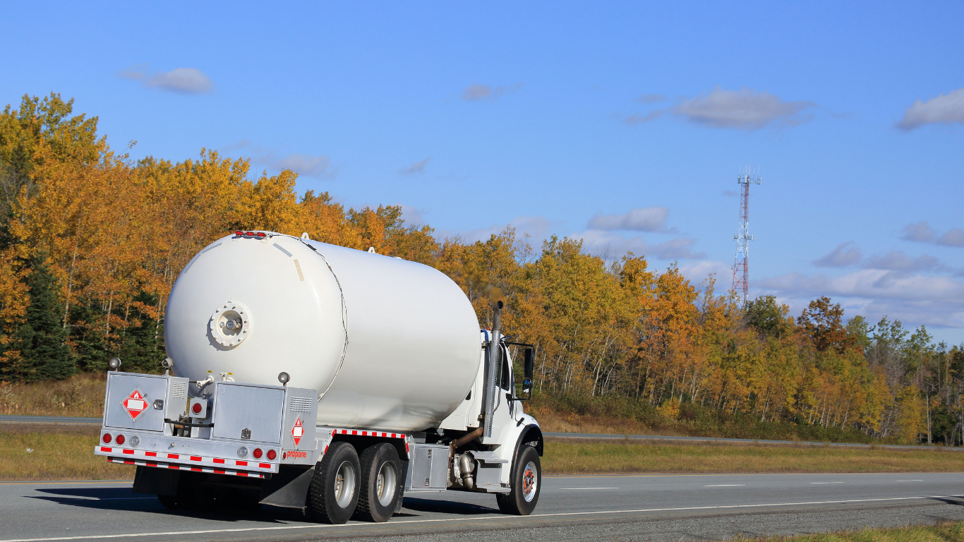 White tanker truck on a highway, with autumn trees and a blue sky in the background.