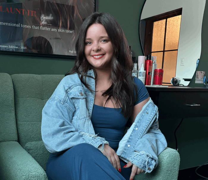 Woman in blue dress and denim jacket smiles, seated on a green couch in front of a mirror and counter.