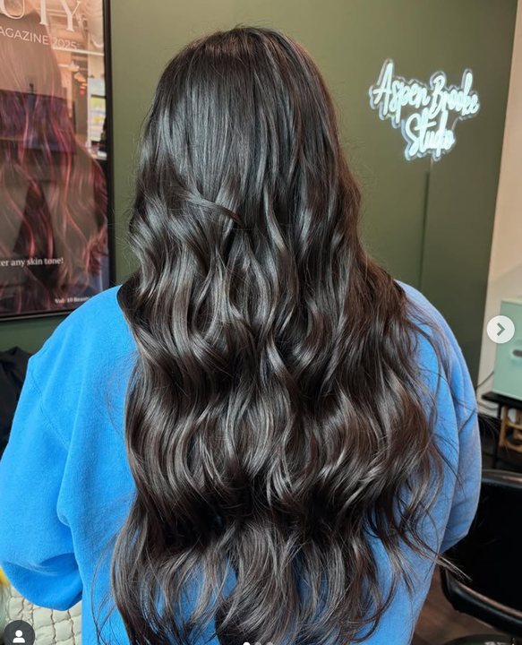 Person from the back with long wavy dark hair in a salon. Blue sweatshirt.