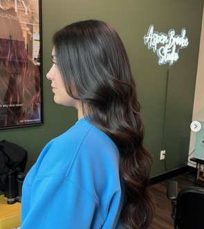 Woman with long, wavy brown hair, wearing a blue sweater, in a hair salon.