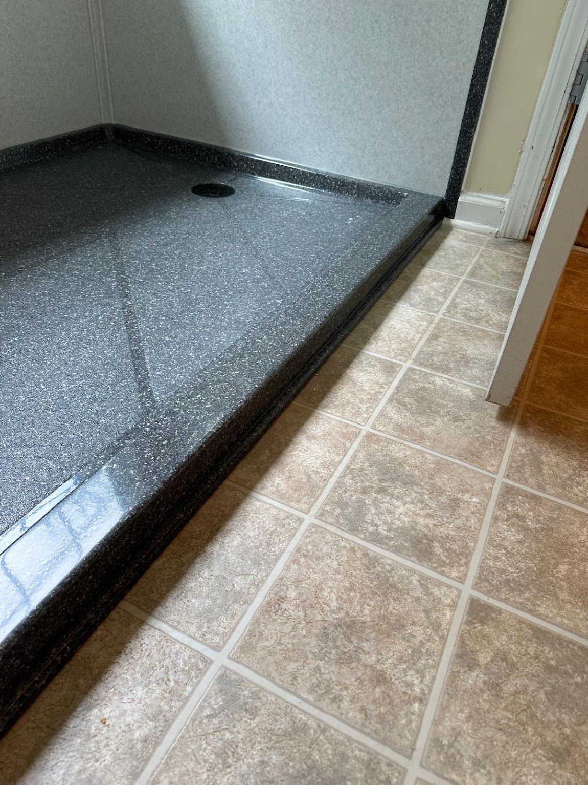 Shower with dark gray base, drain, and tan tiled floor. Light and shadows.