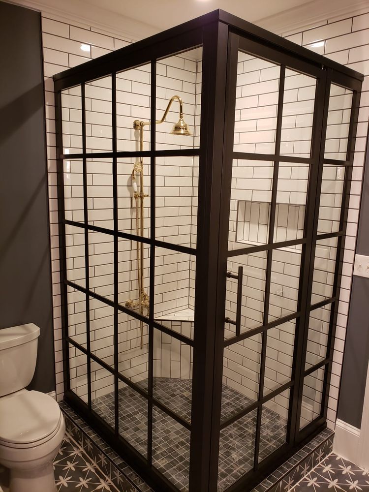 Black-framed glass shower enclosure with white subway tile walls and gold fixtures. The shower floor is tiled in a pattern