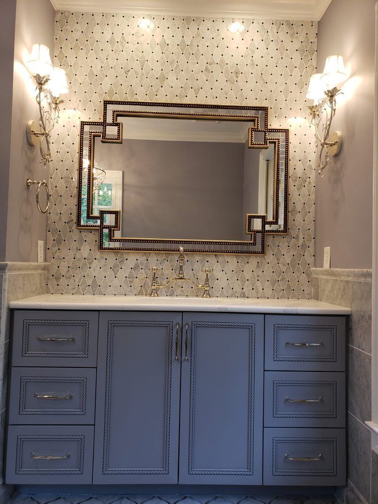 Bathroom vanity with gray cabinets, ornate mirror, patterned wallpaper, and sconce lighting.