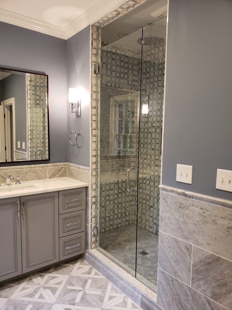 Bathroom with gray cabinets, marble countertops, glass shower, and patterned floor.
