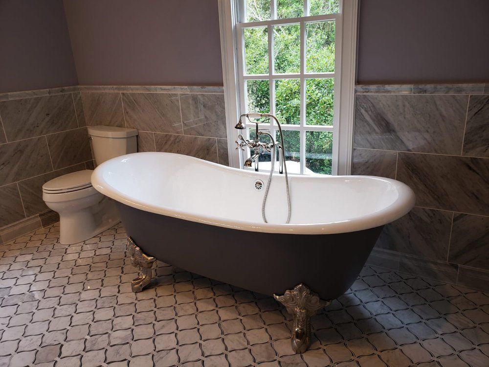 Clawfoot bathtub in a bathroom with window, toilet, and patterned tile floor.