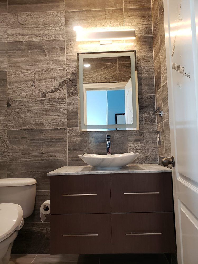 Bathroom with gray tiled walls, brown vanity with sink, mirror with light fixture, and toilet.