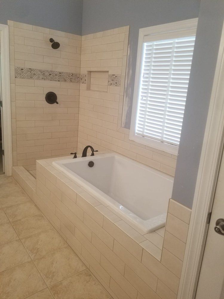 Bathroom with a white bathtub, shower, and light beige tiles. A window with blinds is on the right.