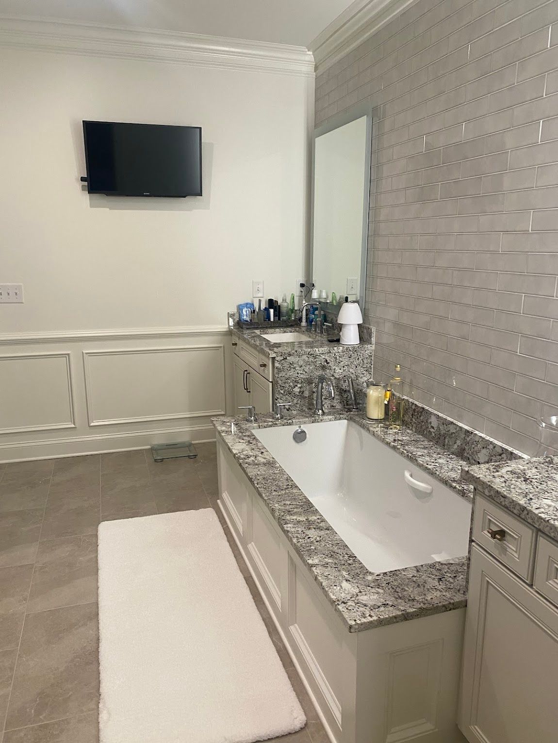 Bathroom with a bathtub, sink, mirror, and TV on the wall. Light-colored walls and countertops.