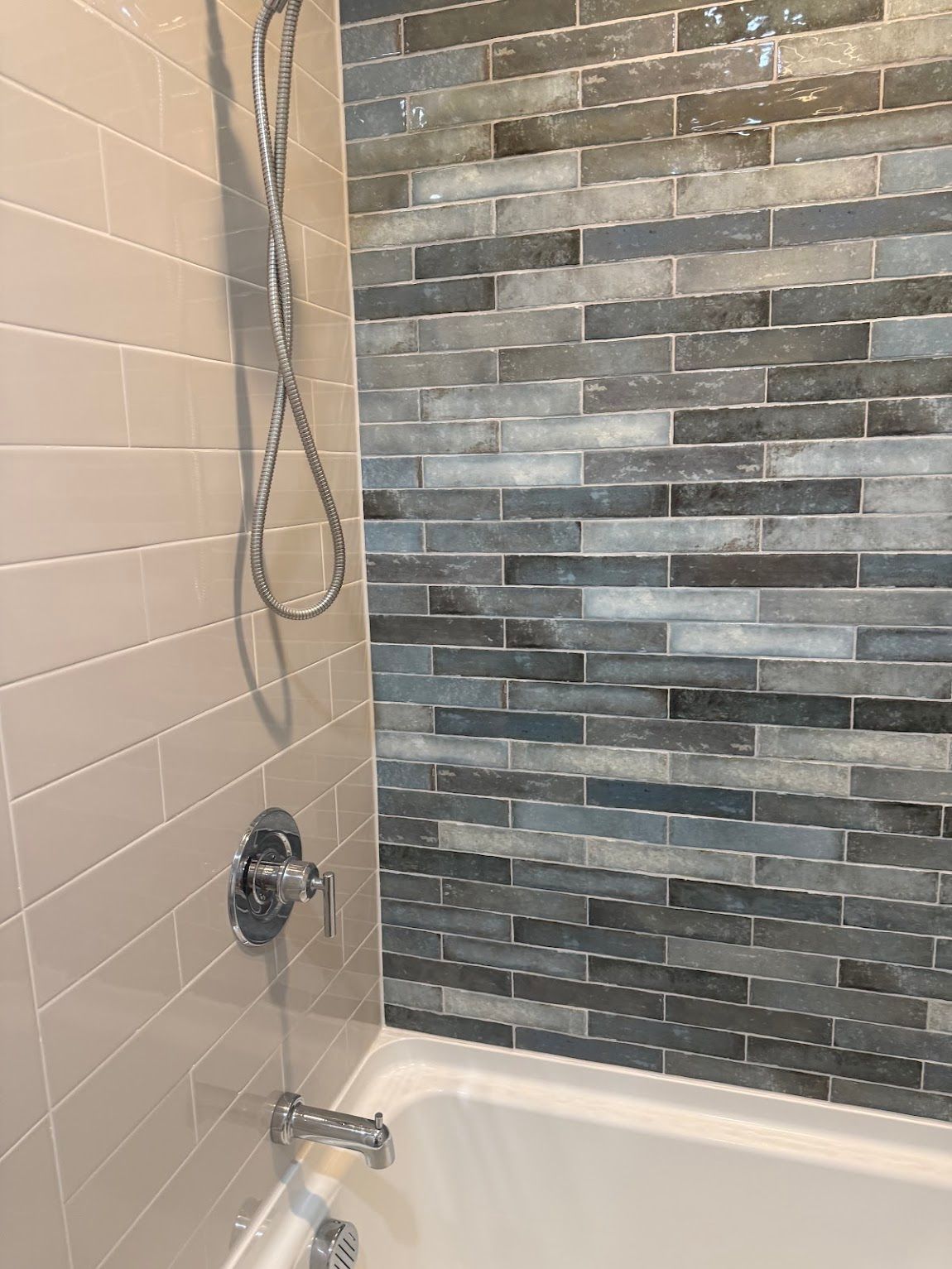 Shower stall with beige and gray tiled walls, chrome fixtures, and a white bathtub.