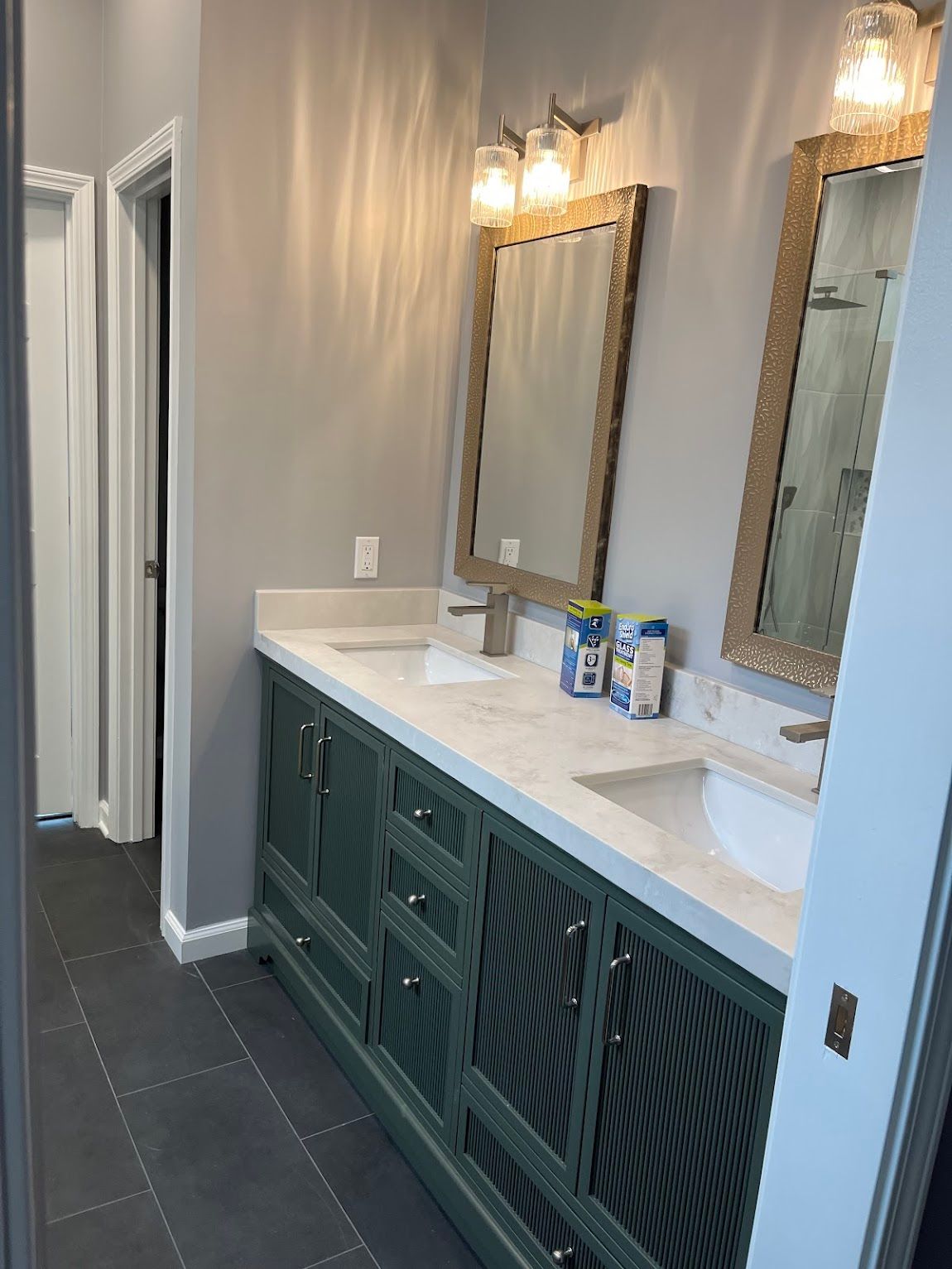 Bathroom with green vanity, two sinks, mirrors, and gray walls. Dark gray tile floor and a door on the left.