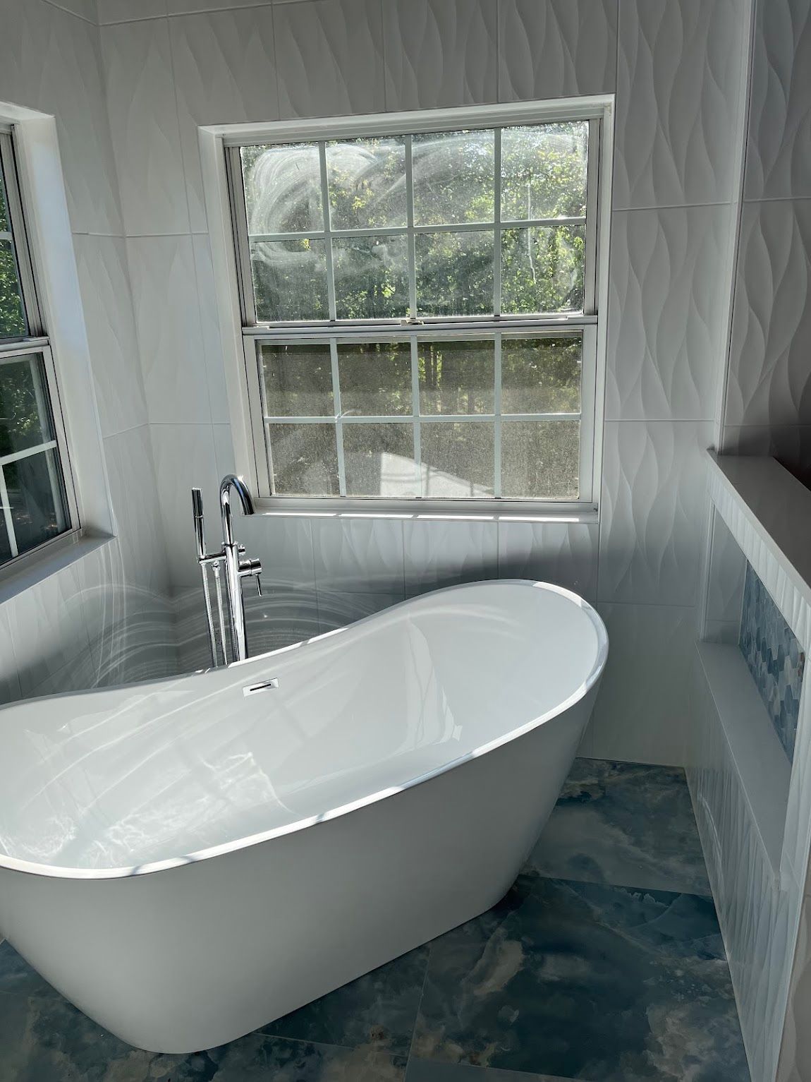 White freestanding bathtub in a modern bathroom with textured white tiles and a window overlooking greenery.