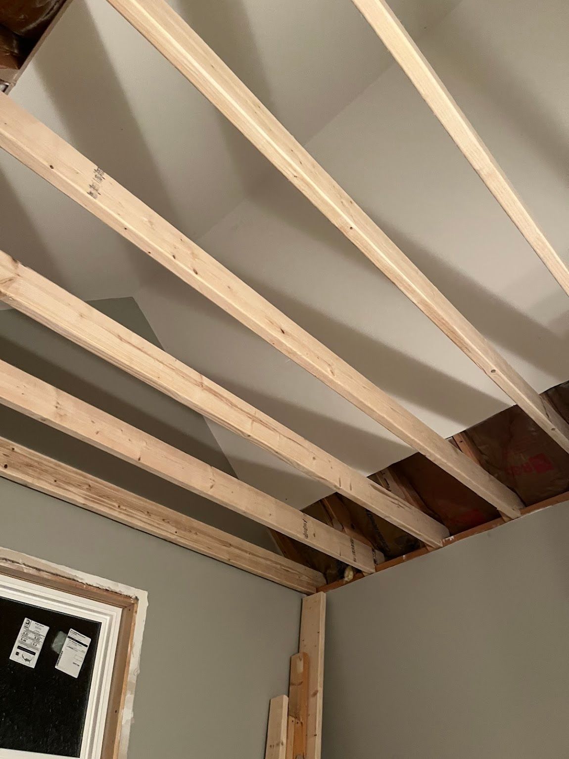 Interior view of a room with exposed ceiling joists. Gray walls and a window frame are visible.